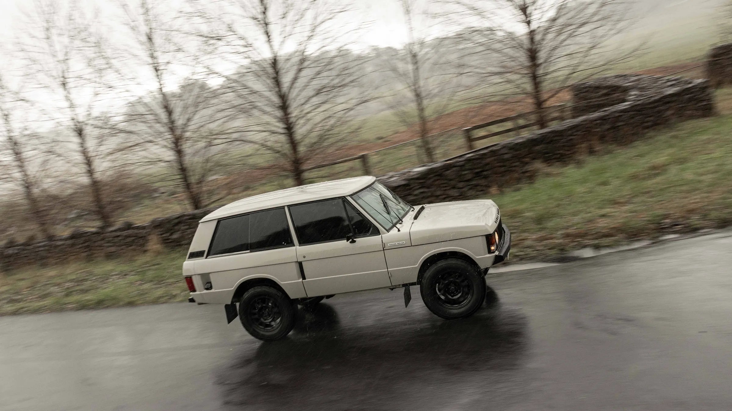 Tan Range Rover driving on a wet road with bare trees and a stone wall in the background | Hudson Coachworks Signature series Range Rover Classic | Range Rover Classic | Range Rover Coachworks | Range Rover Restorations | Range Rover Build | LS Range Rover | V8 Range Rover | Supercharged Range Rover